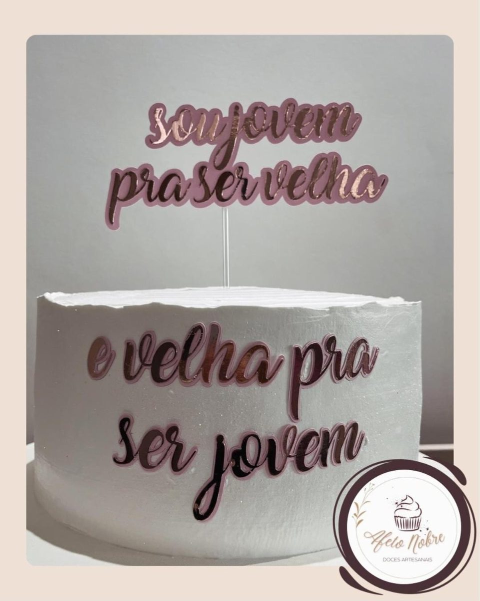 Bolo branco com a frase 'sou jovem pra ser velha e velha pra ser jovem' em topo rosé espelhado